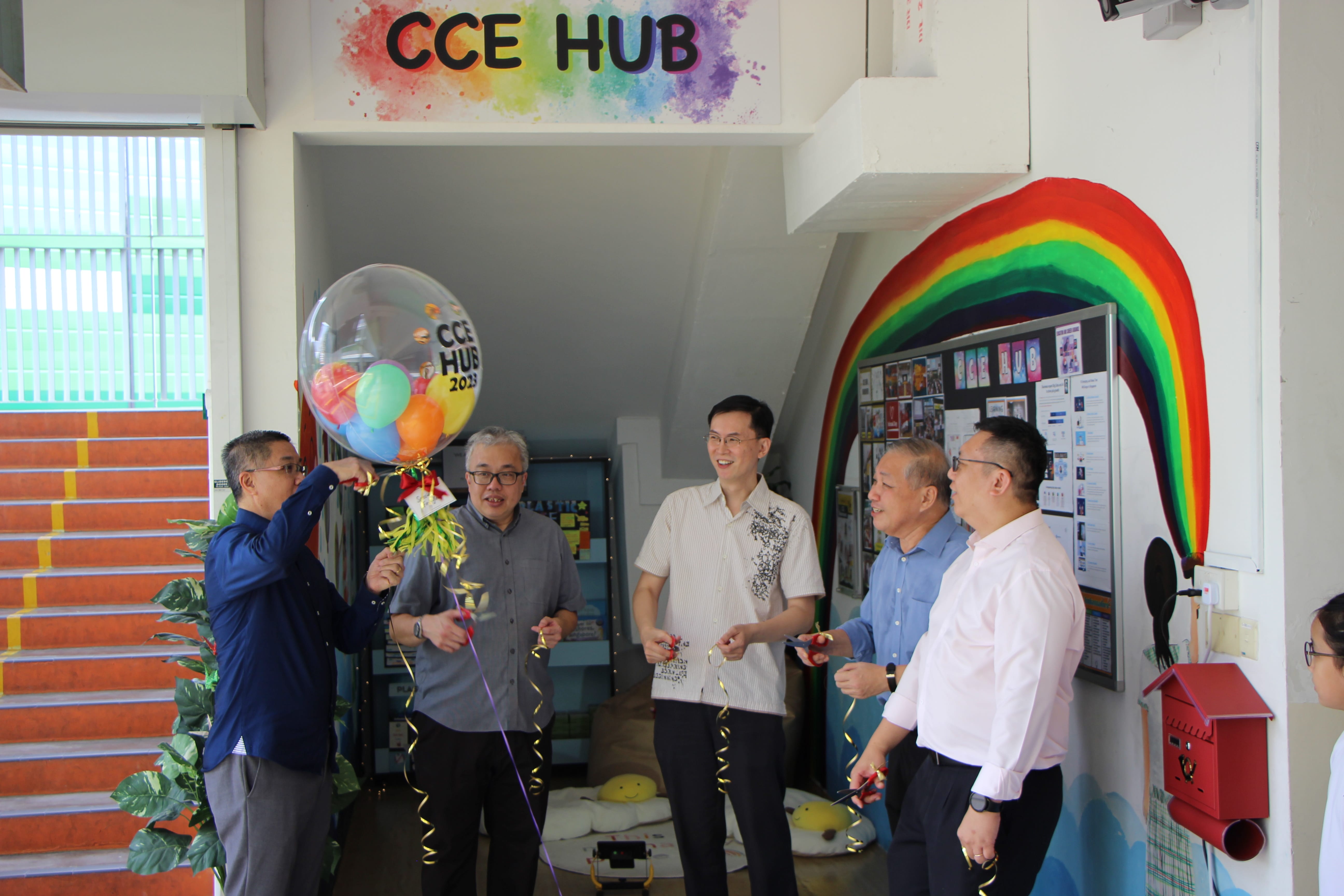 Five men stand near a CCE Hub entrance with a large balloon and rainbow wall decoration during an opening event.